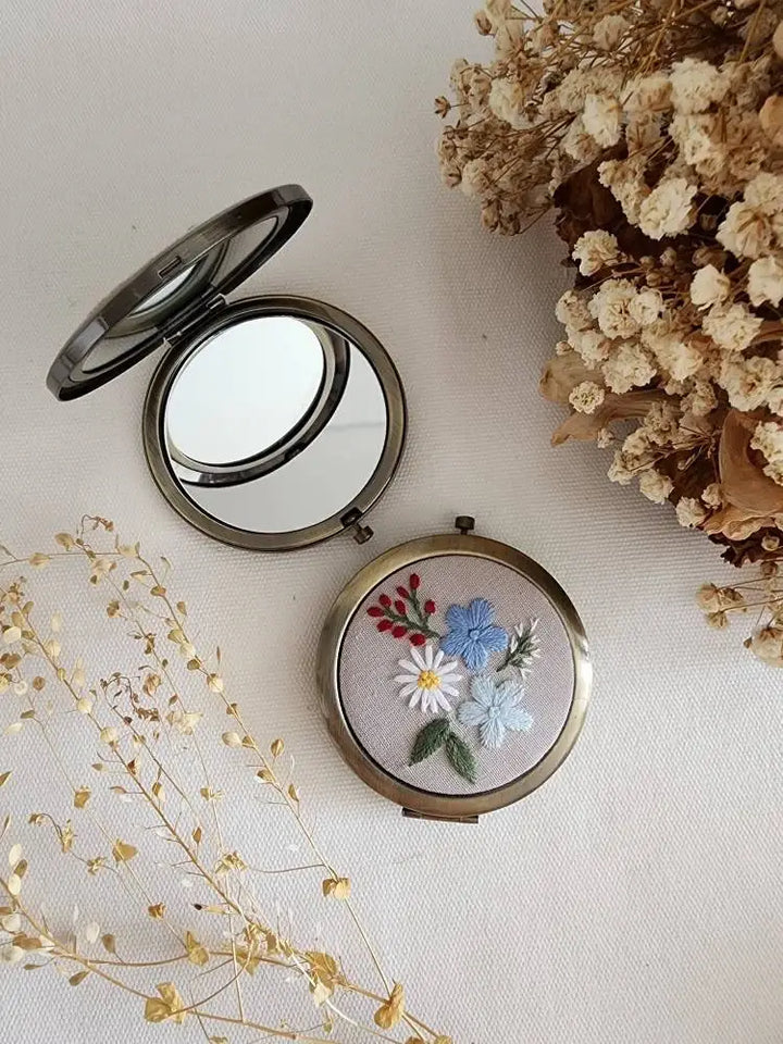 Compact mirror with floral embroidery on a light background with dried flowers.