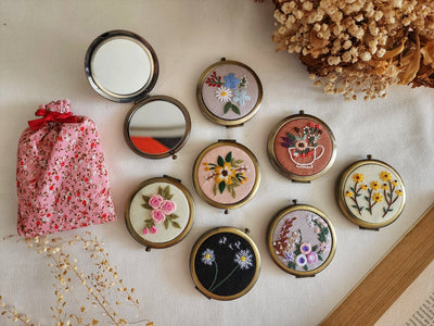 Set of compact mirrors with floral designs on a light surface with a pink floral bag and dried flowers.
